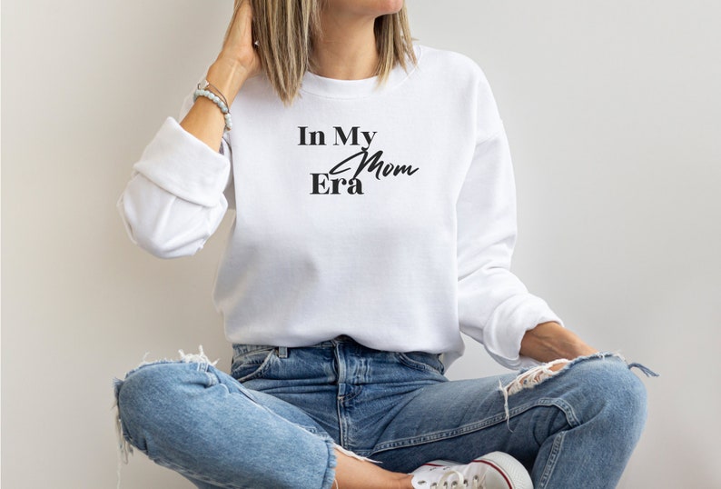 May include: White sweatshirt with the text "In My Mom Era" in black script. The long-sleeved top is paired with blue jeans. The model is wearing white sneakers.