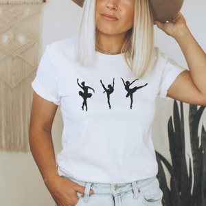 K&ouml;nnte beinhalten: Wei&szlig;es T-Shirt mit einem schwarzen Silhouette-Design von drei Ballettt&auml;nzerinnen in verschiedenen Posen. Die T&auml;nzerinnen werden in Bewegung dargestellt, mit ausgestreckten Armen und Beinen. Das T-Shirt hat einen Rundhalsausschnitt.