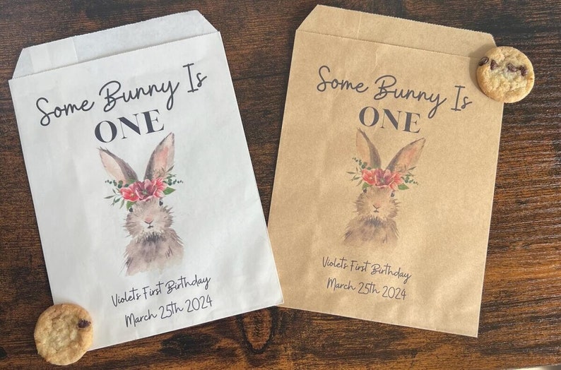 以下が含まれることがあります： 水彩画のウサギのデザインと「Some Bunny Is ONE」の文字が入った、白と茶色の2つの紙袋。袋はバイオレットの1歳の誕生日、2024年3月25日用です。チョコレートチップクッキーが各袋の近くにあります。