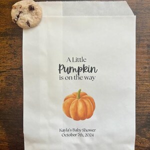 May include: White paper bag with the text "A Little Pumpkin is on the way" and a watercolor illustration of an orange pumpkin. The bag is personalized with "Kayla's Baby Shower October 7th, 2024". A chocolate chip cookie rests on the bag.