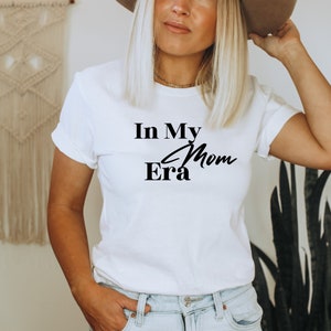 May include: White t-shirt with the text "In My Mom Era" in black script and block letters. The shirt is a crew neck style and is being worn by a person. The background includes a macrame wall hanging and a tall green plant.