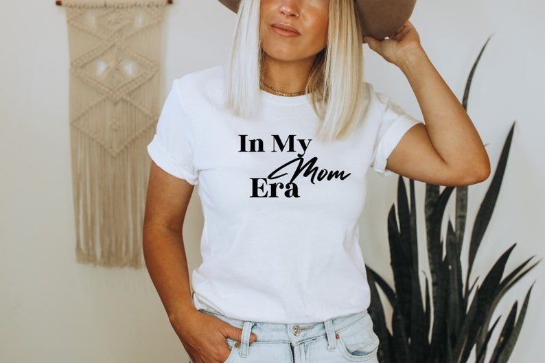 May include: White t-shirt with the text "In My Mom Era" in black script and block letters. The shirt is a crew neck style and is being worn by a person. The background includes a macrame wall hanging and a tall green plant.