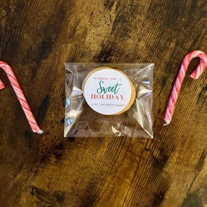 May include: A clear cellophane bag containing a round cookie with a circular sticker that reads "Wishing You a Sweet Holiday" and "Love, The Ippley Family". Two red and white striped candy canes flank the bag on a wooden surface.