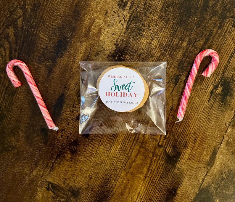 May include: A clear cellophane bag containing a round cookie with a circular sticker that reads "Wishing You a Sweet Holiday" and "Love, The Ippley Family". Two red and white striped candy canes flank the bag on a wooden surface.