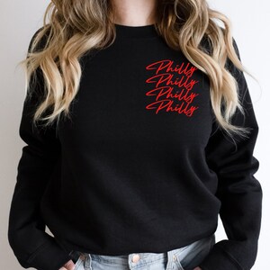 May include: Black crew neck sweatshirt with the word "Philly" in red script repeated three times on the upper left chest. The sweatshirt is a solid black color.