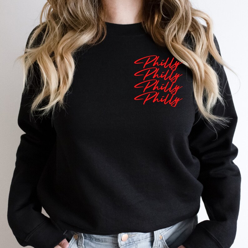May include: Black crew neck sweatshirt with the word "Philly" in red script repeated three times on the upper left chest. The sweatshirt is a solid black color.