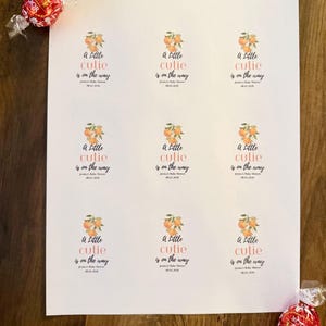 May include: A white sheet featuring nine identical rectangular stickers with an orange fruit illustration and the text A Little cutie is on the way, Jessica's Baby Shower, 08.02.2026. Two red Lindor chocolate truffles are placed at the corners.