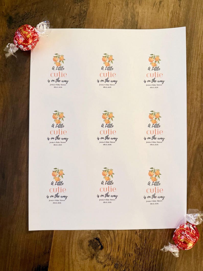 May include: A white sheet featuring nine identical rectangular stickers with an orange fruit illustration and the text A Little cutie is on the way, Jessica's Baby Shower, 08.02.2026. Two red Lindor chocolate truffles are placed at the corners.