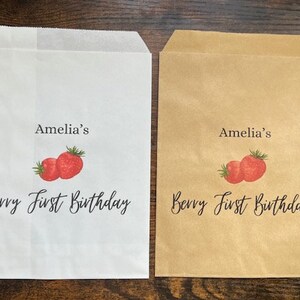 May include: Two paper treat bags, one white and one tan, with a strawberry design. The text "Amelia's Berry First Birthday" is printed on each bag in black script, making them ideal for a first birthday celebration.