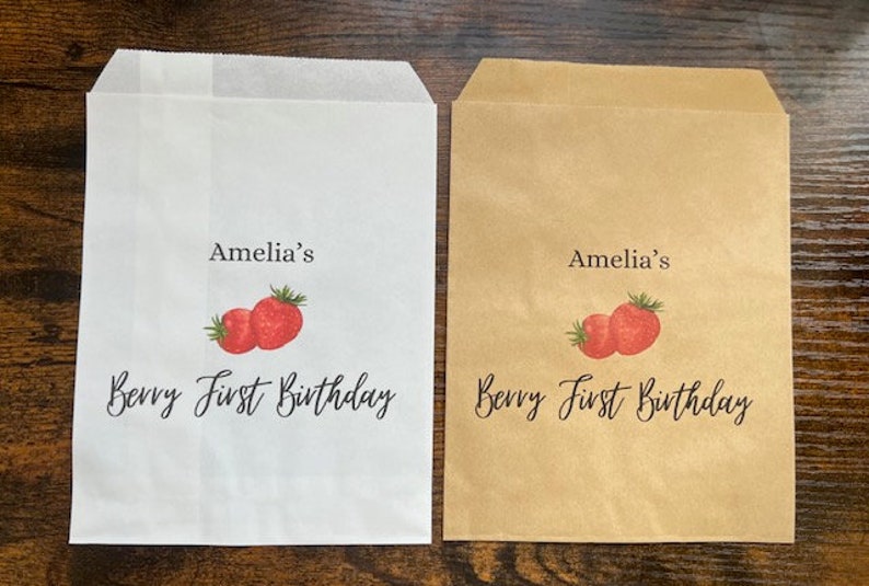 May include: Two paper treat bags, one white and one tan, with a strawberry design. The text "Amelia's Berry First Birthday" is printed on each bag in black script, making them ideal for a first birthday celebration.