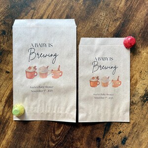 May include: Two brown paper bags with the text "A Baby Is Brewing" and illustrations of coffee cups. The bags also include the text "Kayla's Baby Shower November 5th, 2025". A red and a green decorative item are on the wooden surface.