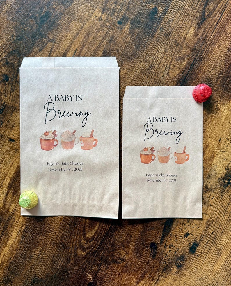 May include: Two brown paper bags with the text "A Baby Is Brewing" and illustrations of coffee cups. The bags also include the text "Kayla's Baby Shower November 5th, 2025". A red and a green decorative item are on the wooden surface.