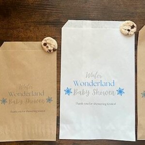 Pu&ograve; includere: Quattro sacchetti di carta, due bianchi e due marroni, con il testo "Winter Wonderland Baby Shower" e grafiche di fiocchi di neve blu. Ogni sacchetto &egrave; sormontato da un biscotto con gocce di cioccolato. Disposti su una superficie di legno scuro.