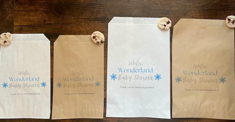 Pu&ograve; includere: Quattro sacchetti di carta, due bianchi e due marroni, con il testo "Winter Wonderland Baby Shower" e grafiche di fiocchi di neve blu. Ogni sacchetto &egrave; sormontato da un biscotto con gocce di cioccolato. Disposti su una superficie di legno scuro.