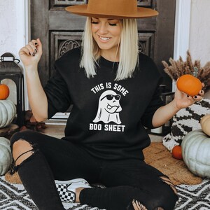 May include: Black sweatshirt with the text "THIS IS SOME BOO SHEET" and a ghost wearing sunglasses. The image includes pumpkins and fall decor, creating a Halloween theme. The person is wearing black jeans and a brown hat.