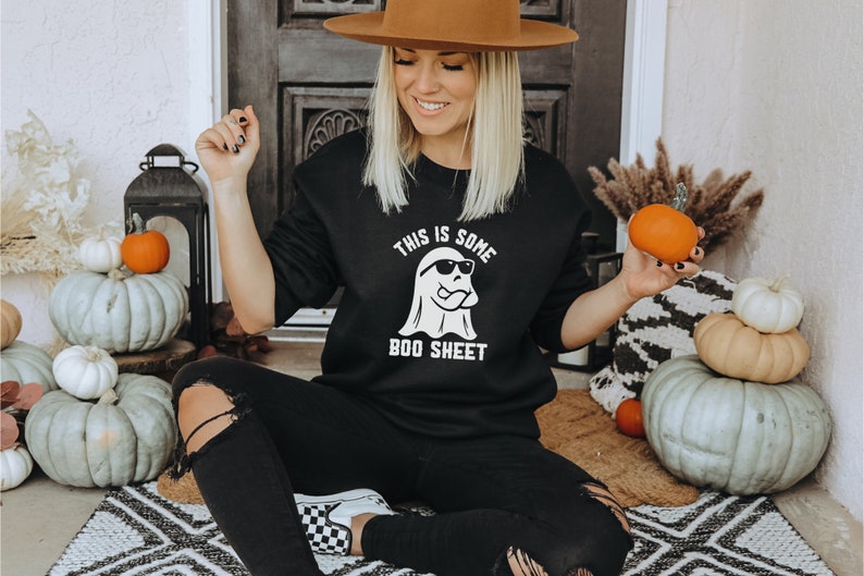 May include: Black sweatshirt with the text "THIS IS SOME BOO SHEET" and a ghost wearing sunglasses. The image includes pumpkins and fall decor, creating a Halloween theme. The person is wearing black jeans and a brown hat.