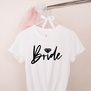 Puede incluir: Camiseta blanca con la palabra "Bride" en fuente negra, coronada por un gr&aacute;fico de diamante. La camiseta est&aacute; colgada de una percha blanca adornada con perlas, con un accesorio de tul rosa.