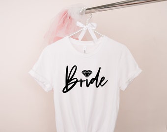 Peut inclure: T-shirt blanc avec le mot "Bride" en noir, surmonté d'un diamant. Le t-shirt est suspendu à un cintre blanc orné de perles, avec un accessoire en tulle rose.