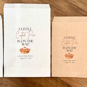 May include: Four rectangular paper bags in white and kraft brown, each printed with the text A LITTLE Cutie Pie IS ON THE WAY!, a small pie illustration, and the text Jessica's Baby Shower August 29th, 2026. A single chocolate cookie is visible.