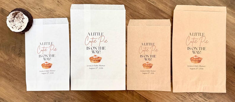 May include: Four rectangular paper bags in white and kraft brown, each printed with the text A LITTLE Cutie Pie IS ON THE WAY!, a small pie illustration, and the text Jessica's Baby Shower August 29th, 2026. A single chocolate cookie is visible.