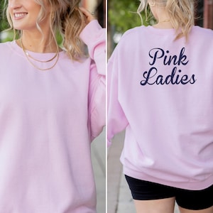 Könnte beinhalten: Hellrosa Sweatshirt mit dem marineblauen Schriftzug "Pink Ladies" auf dem Rücken. Das Sweatshirt mit Rundhalsausschnitt ist einfarbig. Das Model trägt eine goldene Halskette. Das Bild zeigt die Vorder- und Rückseite des Kleidungsstücks.