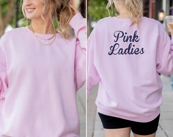 May include: A light pink sweatshirt with the text "Pink Ladies" in navy blue on the back. The sweatshirt is a solid color and has a crew neck. The model is wearing a gold necklace.