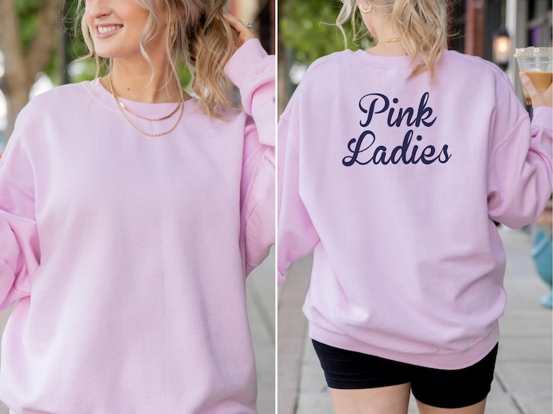 May include: A light pink sweatshirt with the text "Pink Ladies" in navy blue on the back. The sweatshirt is a solid color and has a crew neck. The model is wearing a gold necklace.