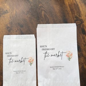 May include: Two white paper bags with the text "SHE'S FRESH OFF the market" in black script. Below the text is "Jessica's Bridal Shower May 10th, 2026". A small floral design is on the right side.