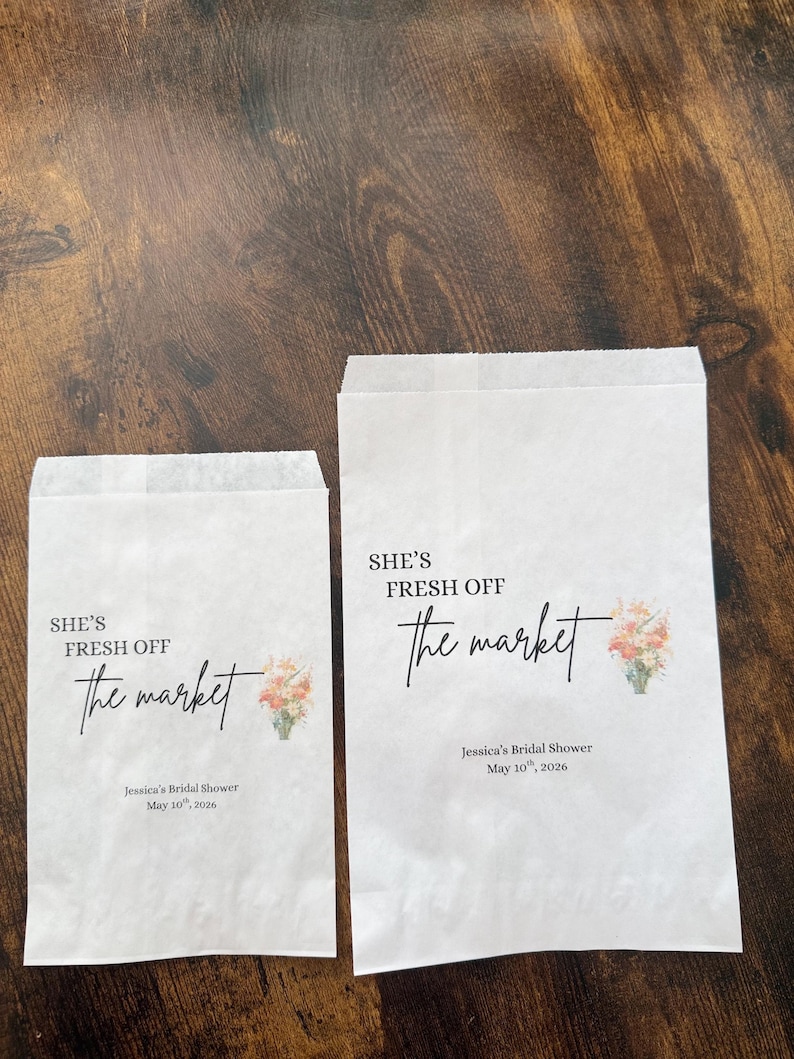 May include: Two white paper bags with the text "SHE'S FRESH OFF the market" in black script. Below the text is "Jessica's Bridal Shower May 10th, 2026". A small floral design is on the right side.