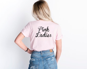 May include: A light pink t-shirt with the words "Pink Ladies" in black script. The shirt is worn by a person with blonde hair and blue jeans. The sleeves are rolled up.