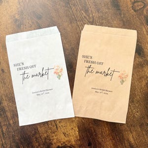 Pode incluir: Duas sacolas de papel para lembrancinhas, uma branca e outra bege, com o texto "SHE'S FRESH OFF the market" em escrita preta. Cada sacola tem uma ilustração floral e o texto "Jessica's Bridal Shower May 10th, 2026".