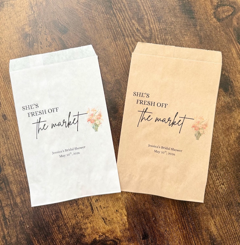 May include: Two paper favor bags, one white and one tan, with the text "SHE'S FRESH OFF the market" in black script. Each bag has a floral illustration and the text "Jessica's Bridal Shower May 10th, 2026".