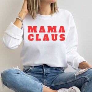Op de afbeelding: Witte sweatshirt met de rode tekst "MAMA CLAUS". De lange mouwen top wordt gecombineerd met een blauwe jeans. De sweatshirt is een casual stijl, perfect voor de feestdagen.