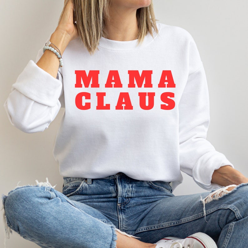 Op de afbeelding: Witte sweatshirt met de rode tekst "MAMA CLAUS". De lange mouwen top wordt gecombineerd met een blauwe jeans. De sweatshirt is een casual stijl, perfect voor de feestdagen.