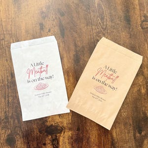 May include: Two paper favor bags, one white and one tan, with the text "A Little Meatball is on the way!" in burgundy script. A plate of spaghetti and meatballs is illustrated below the text. The bags are for Jessica's Baby Shower.