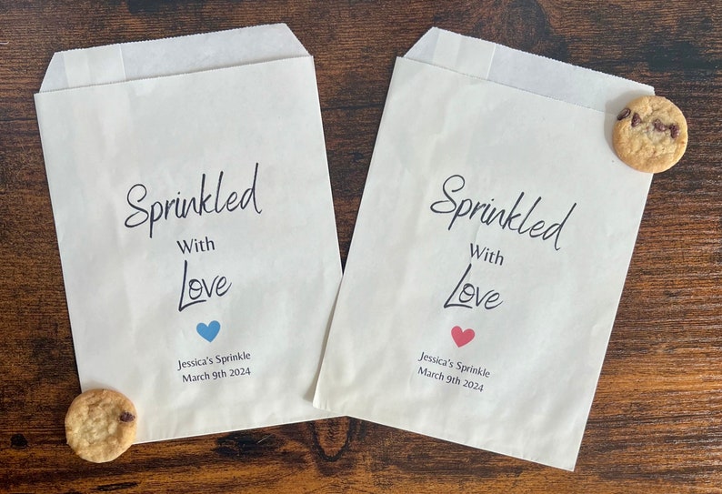 May include: Two white paper treat bags with the text "Sprinkled With Love" in black script. Each bag has a heart or a blue heart below the text, with the text "Jessica's Sprinkle March 9th 2024". Chocolate chip cookies are next to the bags.