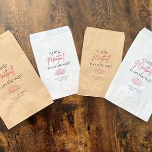 May include: Four paper favor bags, two in brown and two in white, with the text "A Little Meatball is on the way!" in a script font. The bags are for a baby shower.