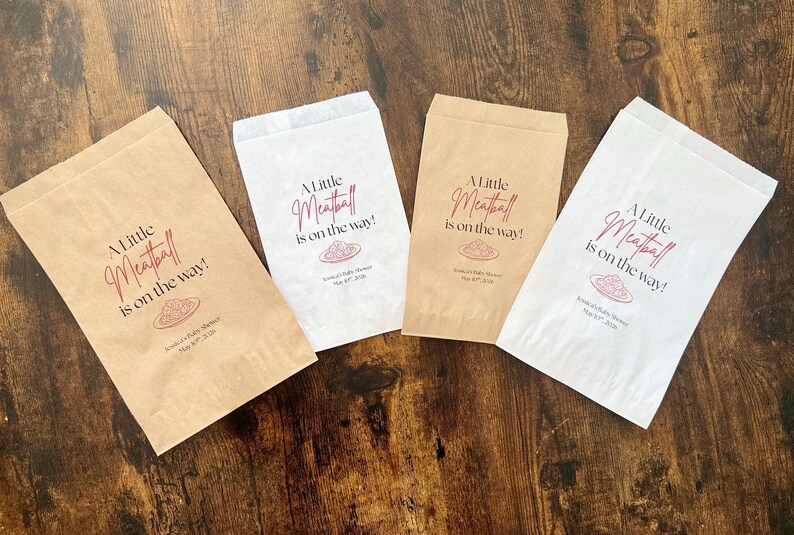 May include: Four paper favor bags, two in brown and two in white, with the text "A Little Meatball is on the way!" in a script font. The bags are for a baby shower.