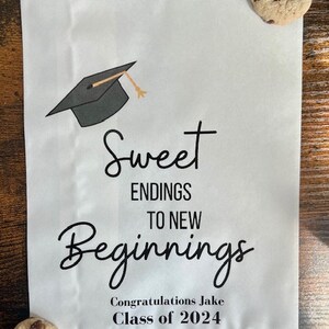 May include: White paper bag with a graduation cap graphic and the text "Sweet Endings to New Beginnings." The bag also includes the text "Congratulations Jake Class of 2024." Two chocolate chip cookies are placed on the bag.