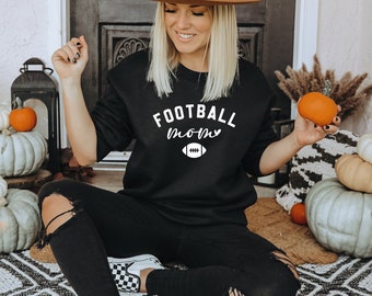 Może przedstawiać: Czarna bluza z napisem "FOOTBALL mom" i grafiką piłki nożnej. Bluza jest połączona z czarnymi dżinsami. Obraz ma jesienny motyw z dyniami i ozdobnymi tykwami.