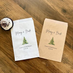 May include: Two rectangular paper favor bags, one white and one kraft brown, each printed with a green watercolor pine tree, the text Merry & Bride, and the personalized details Jessica's Bridal Shower 12.20.26, alongside a small chocolate treat.
