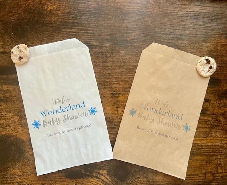 Pu&ograve; includere: Due sacchetti di carta, uno bianco e uno marrone, con la scritta "Winter Wonderland Baby Showen" in blu e grigio. Ogni sacchetto ha un biscotto con gocce di cioccolato attaccato. Il testo "Thank you for showering Jessica!" &egrave; stampato anche sui sacchetti.