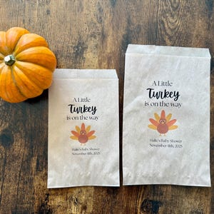 May include: An orange pumpkin next to two paper bags with the text "A Little Turkey is on the way". The bags also feature a turkey graphic and the text "Halle's Baby Shower November 11th, 2025".