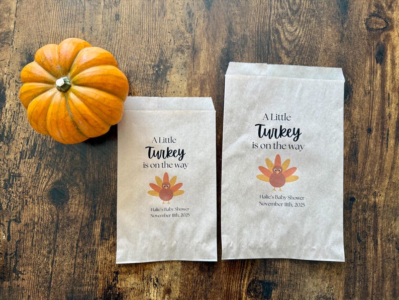 May include: An orange pumpkin next to two paper bags with the text "A Little Turkey is on the way". The bags also feature a turkey graphic and the text "Halle's Baby Shower November 11th, 2025".