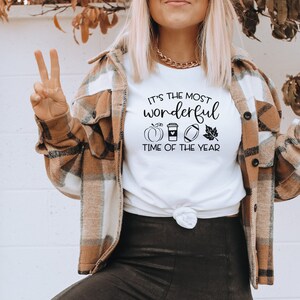 Puede incluir: Camiseta blanca con el texto "It's the most wonderful time of the year" y gr&aacute;ficos tem&aacute;ticos de oto&ntilde;o. La camiseta se combina con una chaqueta a cuadros marr&oacute;n y crema. La modelo lleva un collar de cadena dorada.