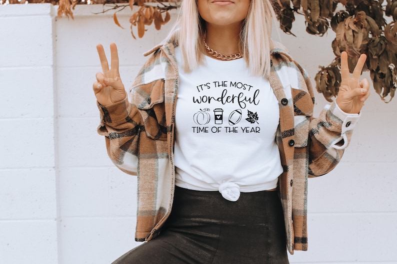 Puede incluir: Camiseta blanca con el texto "It's the most wonderful time of the year" y gr&aacute;ficos tem&aacute;ticos de oto&ntilde;o. La camiseta se combina con una chaqueta a cuadros marr&oacute;n y crema. La modelo lleva un collar de cadena dorada.