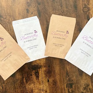Puede incluir: Cuatro bolsas de papel para golosinas con el texto "Our Little Butterfly is Turning One!" en letra cursiva, con un gr&aacute;fico de mariposa. Las bolsas son en tonos de blanco y marr&oacute;n, dispuestas sobre una superficie de madera oscura.