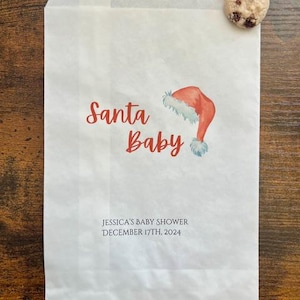 May include: White paper bag printed with "Santa Baby" in red script and a watercolour Santa hat illustration. The bag also features the text "JESSICA'S BABY SHOWER DECEMBER 17TH, 2024". A chocolate chip cookie is placed on the top right corner.