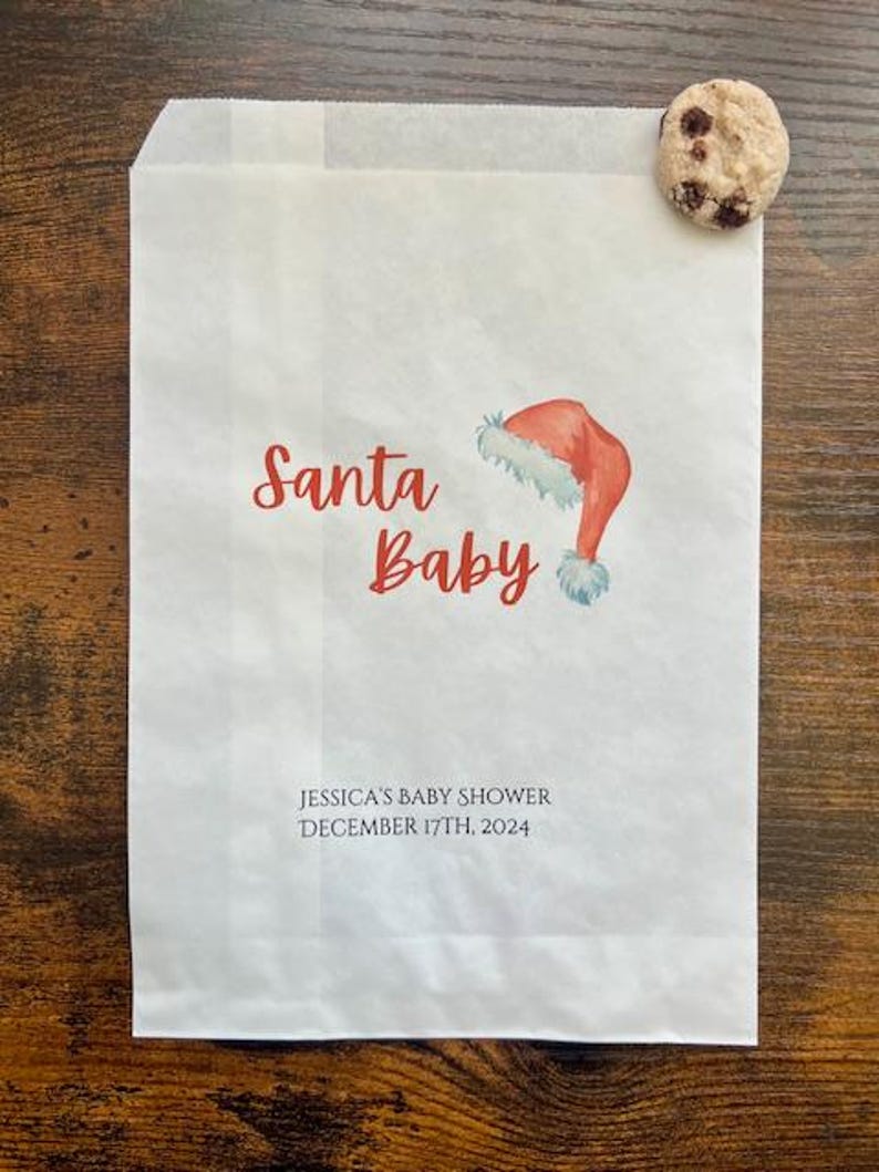 May include: White paper bag printed with "Santa Baby" in red script and a watercolour Santa hat illustration. The bag also features the text "JESSICA'S BABY SHOWER DECEMBER 17TH, 2024". A chocolate chip cookie is placed on the top right corner.