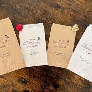 May include: Four paper favor bags with the text "A Little Butterfly is on the Way!" in a script font, with a butterfly graphic. Each bag has a different shade of brown or white. A red or gold foil-wrapped chocolate is attached to each bag.